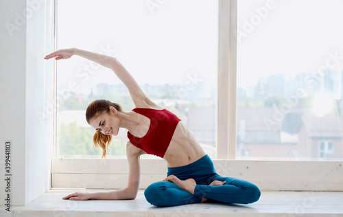 A sportive woman in jeans and a tank top practices yoga near the window