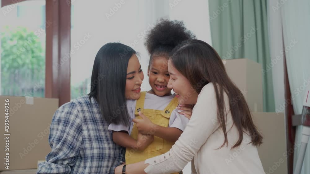 A young happy Asian lesbian couple sitting on the floor with cardboard boxes hugging their lovely African daughter standing in the middle between them, twisting her finger with an embarrassed smile. A young happy Asian lesbian couple sitting on the floor with cardboard boxes hugging their lovely African daughter standing in the middle between them, twisting her finger with an embarrassed smile.