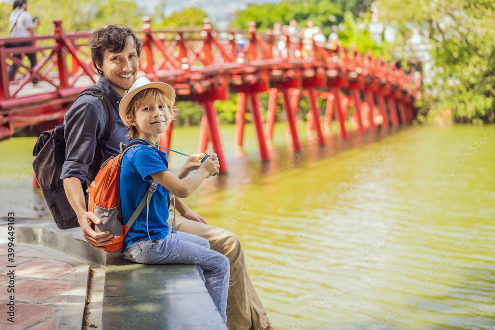 Caucasian Dad and son travelers on background of Red Bridge in public park garden with trees and reflection in the middle of Hoan Kiem Lake in Downtown Hanoi. Traveling with children concept