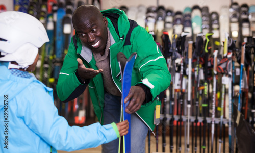 Positive African American man choosing ski for his preteen son in store of sports equipment