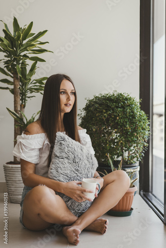 Young European woman with long straight hair sitting on the floor near potted plants, drinking morning coffee and looking through the window. Morning routine, mindfulness or self-care concept