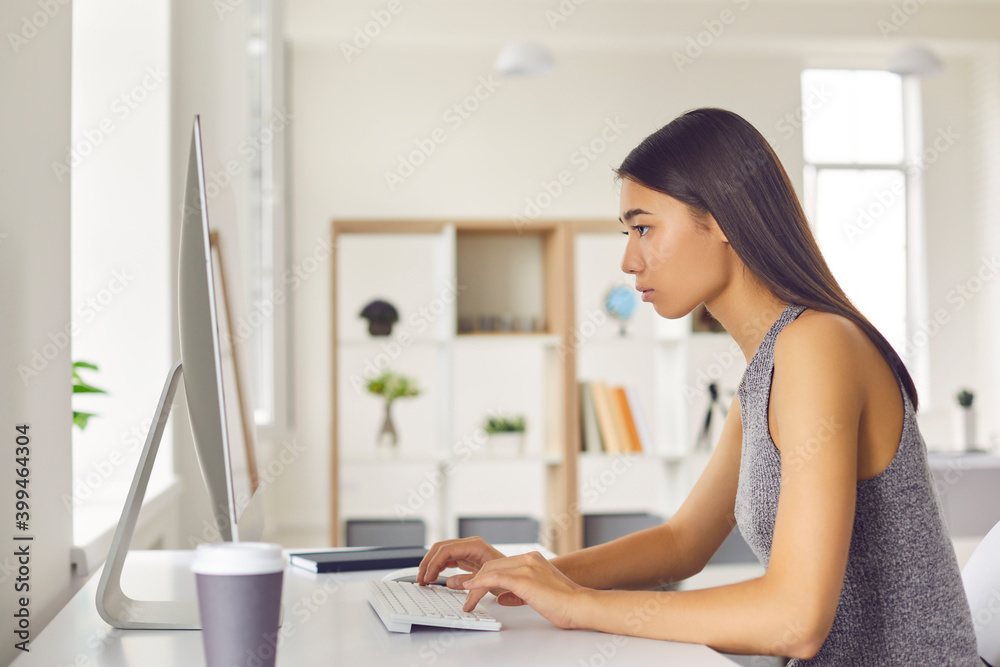 Young pretty asian woman office worker sitting at computer and monitor and working online, side view Young pretty asian woman office worker sitting at computer and monitor and working online, side view