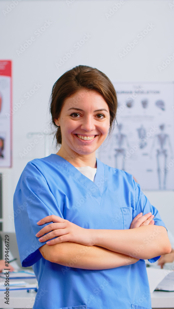 Portrait of young nurse standing in front of camera smiling in medical conference meeting office. Team of specialist doctors working in background talking about symptoms of disease in clinic room