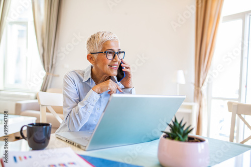 Focused old woman with white hair at home making a business call wile using laptop. Senior stylish entrepreneur wearing eyeglasses working at home. Woman managing domestic bills and home finance photo