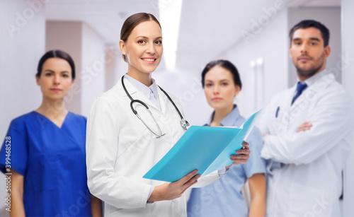 medicine  profession and healthcare concept - happy smiling female doctor in white coat with folder and stethoscope over group of colleagues at hospital on background