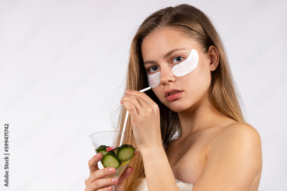 Blonde with eye patches under the eyes, holding a detox cucumber cocktail. Healthy care. White background. Blonde with eye patches under the eyes, holding a detox cucumber cocktail. Healthy care. White background.