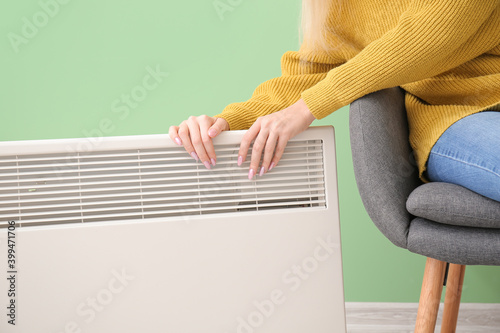 Young woman warming hands near electric heater. Concept of heating season photo