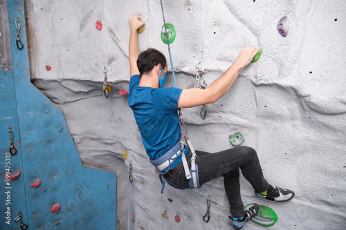 Active sporty man wearing protective face mask practicing rock climbing on artificial rock in a climbing wall. New normal in extreme sports.
