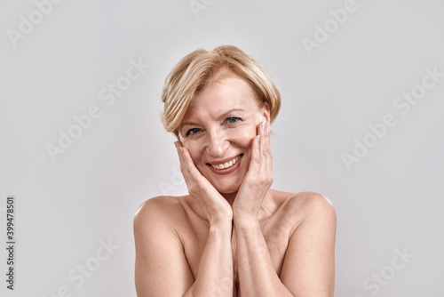 Portrait of beautiful naked middle aged woman looking excited, smiling at camera, touching her cheeks while posing isolated against grey background. Beauty, skincare concept