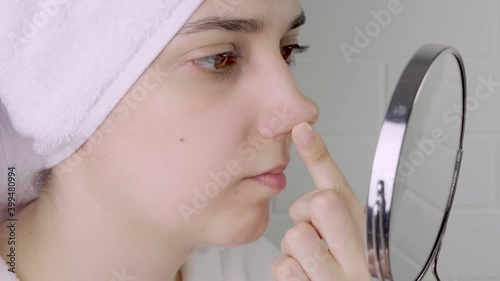 Black dots on the nose. A young woman looks at her face in the mirror. Skin care, problem skin, cleansing. Natural beauty, naturalness. Girl without makeup in the bathroom. Blackheads and comedones.
