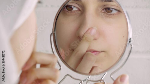 Black dots on the nose. A young woman looks at her face in the mirror. Skin care, problem skin, cleansing. Natural beauty, naturalness. Girl without makeup in the bathroom. Blackheads and comedones.