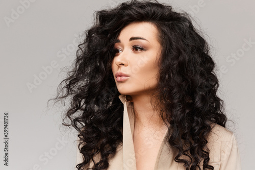 Portrait of the young woman with black curly hair and gentle makeup at the grey background, isolated with copy space