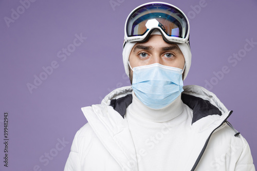 Skier man in warm white windbreaker jacket ski goggles face mask to safe from coronavirus virus covid-19 spend extreme weekend winter in mountains isolated on purple background. People hobby concept.