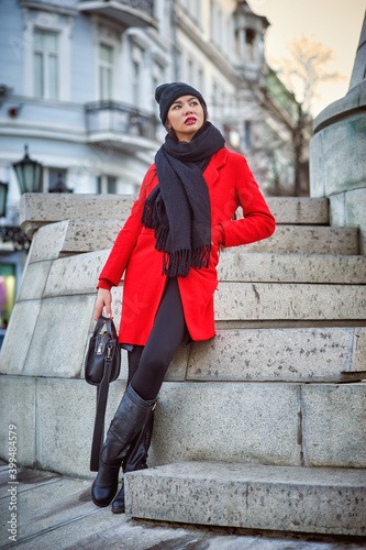 Beautiful young woman of Asian appearance on a winter city street . A girl in a red coat and a knitted hat