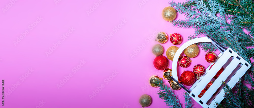 New Year's Christmas tree decorations of red and gold color lie in a chaotic manner on a pink background among fir branches covered with snow, and a wooden box stands nearby. New Year's Christmas tree decorations of red and gold color lie in a chaotic manner on a pink background among fir branches covered with snow, and a wooden box stands nearby.