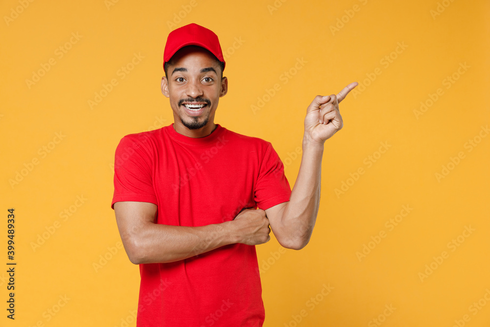 Delivery employee african man in red cap blank tshirt uniform workwear work courier service on quarantine coronavirus covid19 virus concept pointing on free space isolated on yellow background studio