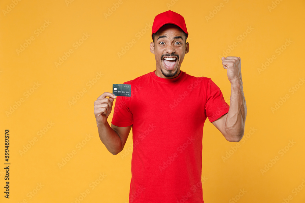 Smiling delivery employee african man 20s in red cap blank print t-shirt uniform workwear work courier dealer service concept hold mockup plastic credit bank card isolated on yellow background studio. Smiling delivery employee african man 20s in red cap blank print t-shirt uniform workwear work courier dealer service concept hold mockup plastic credit bank card isolated on yellow background studio.