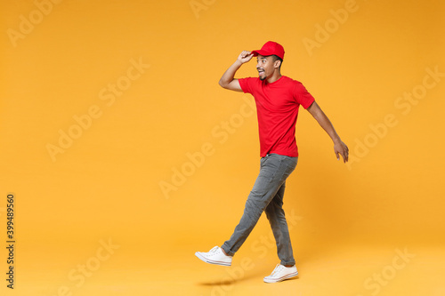 Fototapeta Naklejka Na Ścianę i Meble -  Full length delivery employee african man 20s in red cap blank print t-shirt uniform workwear work courier dealer service during quarantine covid-19 virus concept isolated on yellow background studio.