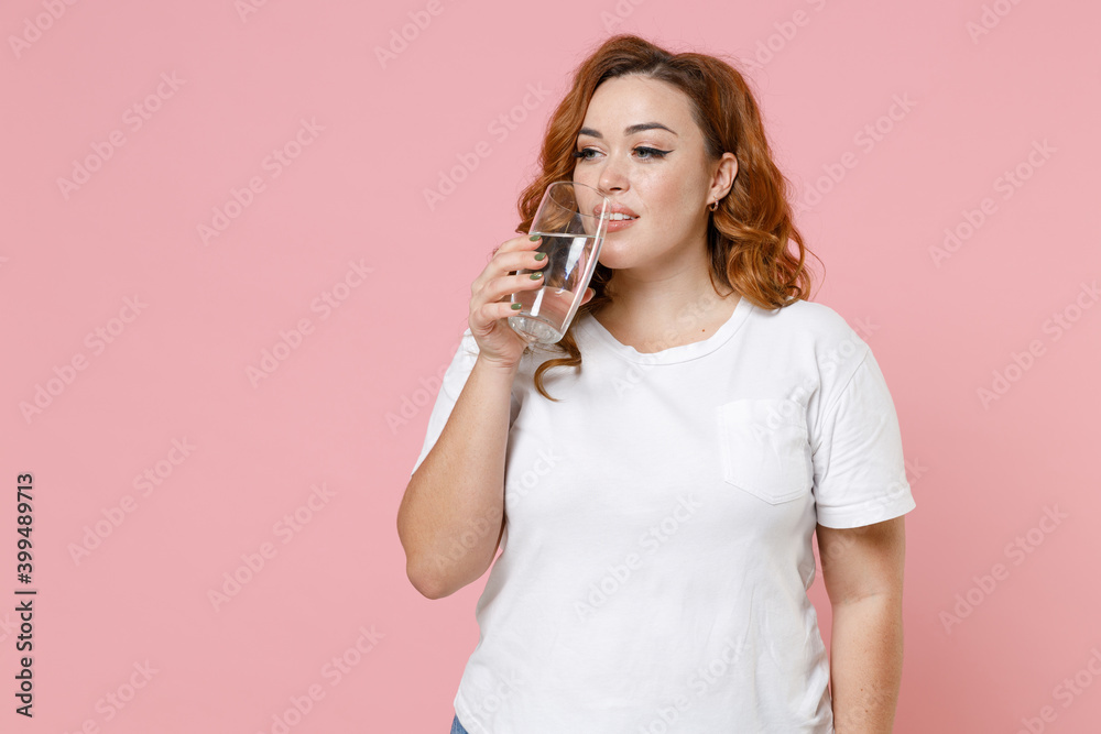 Attractive beautiful young redhead plus size body positive woman 20s wearing white casual t-shirt hold glass of water drinking looking aside isolated on pastel pink color background studio portrait. Attractive beautiful young redhead plus size body positive woman 20s wearing white casual t-shirt hold glass of water drinking looking aside isolated on pastel pink color background studio portrait.