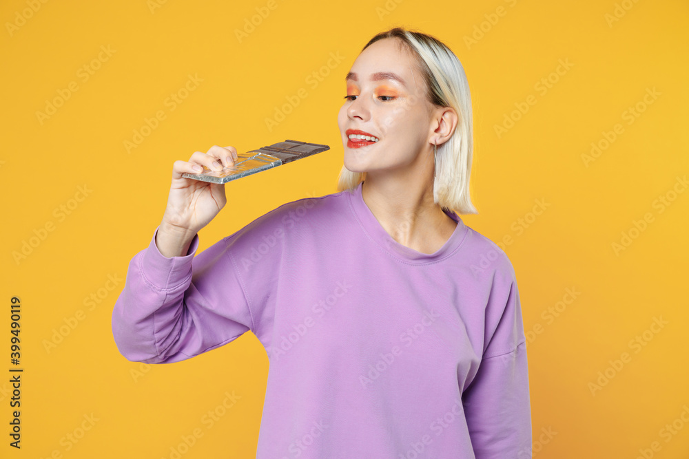 Young blonde smiling caucasian woman 20s with bob haircut bright makeup wearing casual basic purple shirt eating holding in hand chocolate bar isolated on yellow color background studio portrait. Young blonde smiling caucasian woman 20s with bob haircut bright makeup wearing casual basic purple shirt eating holding in hand chocolate bar isolated on yellow color background studio portrait.