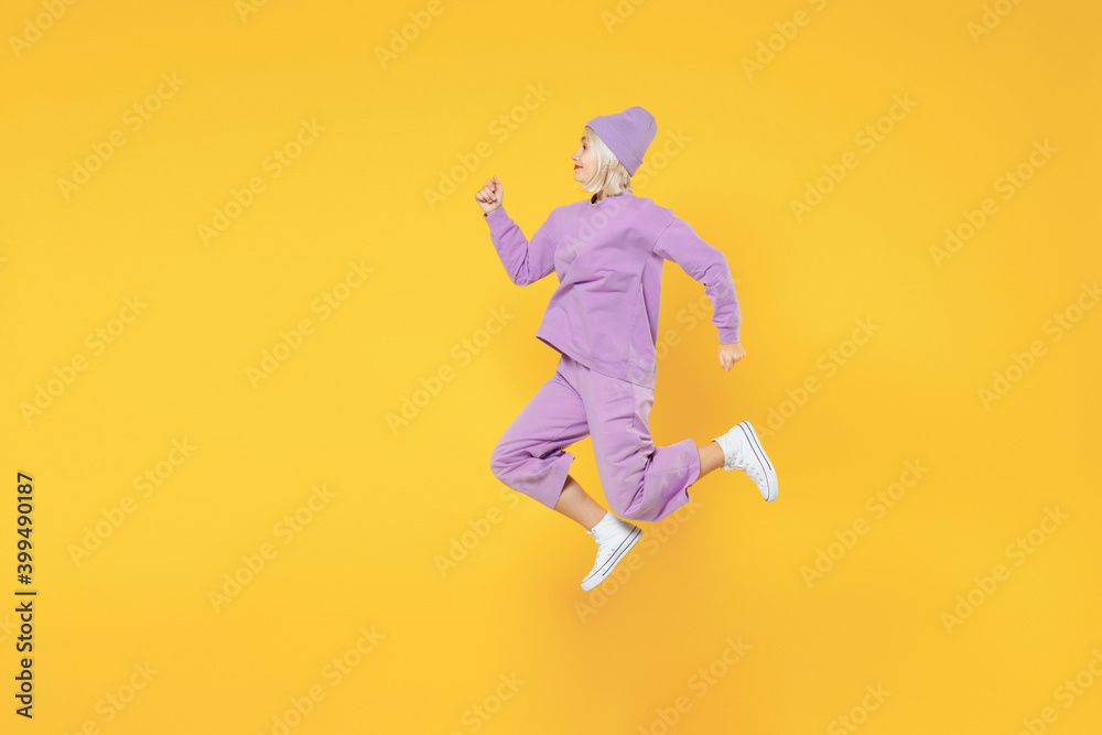 Full length of young energetic caucasian excited fun woman 20s wearing casual basic purple suit beanie hat side profile view running jumping isolated on yellow color background studio portrait