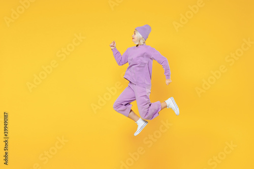 Full length of young energetic caucasian excited fun woman 20s wearing casual basic purple suit beanie hat side profile view running jumping isolated on yellow color background studio portrait