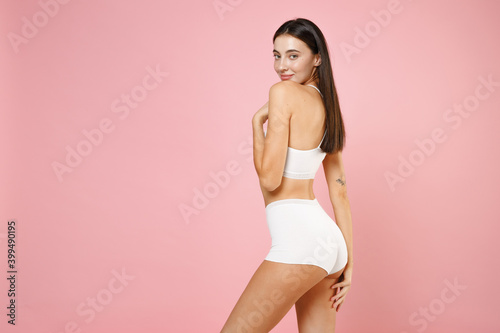 Side view of smiling sexy pretty young brunette woman 20s in white underwear with perfect fit shape posing hold hands on legs looking camera isolated on pastel pink colour background, studio portrait.