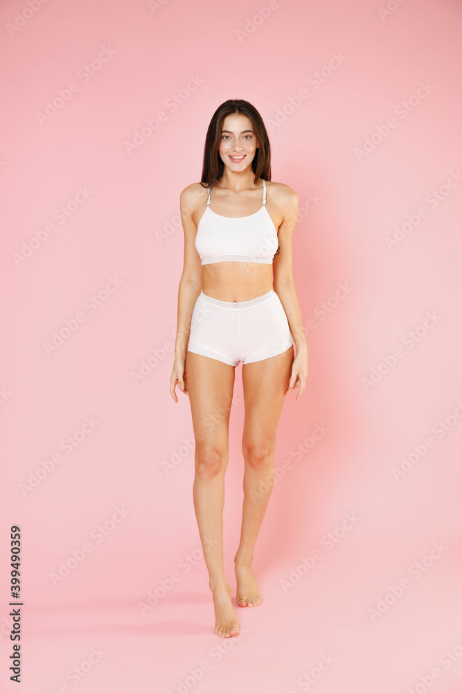 Foto de Full length of smiling beautiful fitness young brunette woman in  white underwear showing fit strong body posing standing looking camera  isolated on pastel pink colour background, studio portrait. do Stock