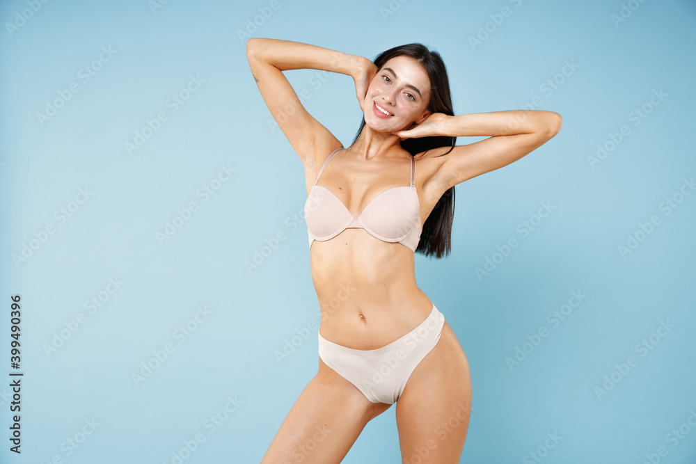 Smiling pretty young brunette woman 20s in beige underwear showing sports body standing posing holding hands behind head looking camera isolated on pastel blue colour wall background studio portrait. Smiling pretty young brunette woman 20s in beige underwear showing sports body standing posing holding hands behind head looking camera isolated on pastel blue colour wall background studio portrait.