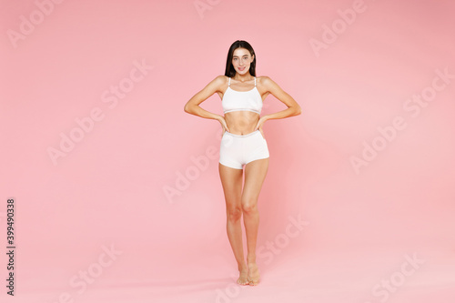 Full length of smiling stunning young brunette woman in white underwear showing fit strong body posing standing with arms akimbo on waist isolated on pastel pink colour background, studio portrait. © ViDi Studio