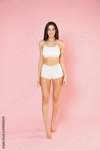Full length of smiling beautiful fitness young brunette woman in white underwear showing fit strong body posing standing looking camera isolated on pastel pink colour background, studio portrait. © ViDi Studio