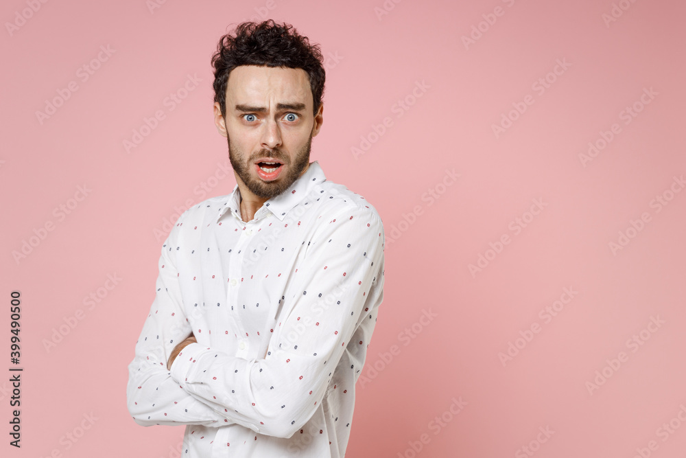 Fototapeta premium Shocked worried scared young bearded man 20s wearing basic casual white shirt standing holding hands crossed keeping mouth open looking camera isolated on pastel pink color background studio portrait.