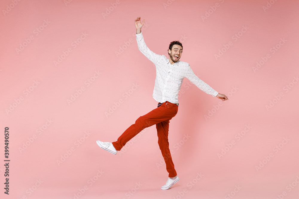 Full length side view of excited young bearded man 20s wearing basic casual white shirt dancing spreading hands rising leg looking camera isolated on pastel pink color wall background studio portrait. Full length side view of excited young bearded man 20s wearing basic casual white shirt dancing spreading hands rising leg looking camera isolated on pastel pink color wall background studio portrait.