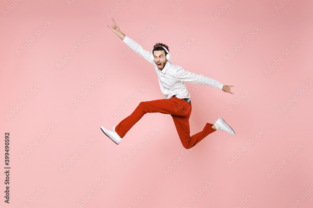 Full length side view crazy young bearded man 20s in white shirt jumping listening music with headphones depicting heavy metal rock sign horns up gesture isolated on pink background studio portrait.
