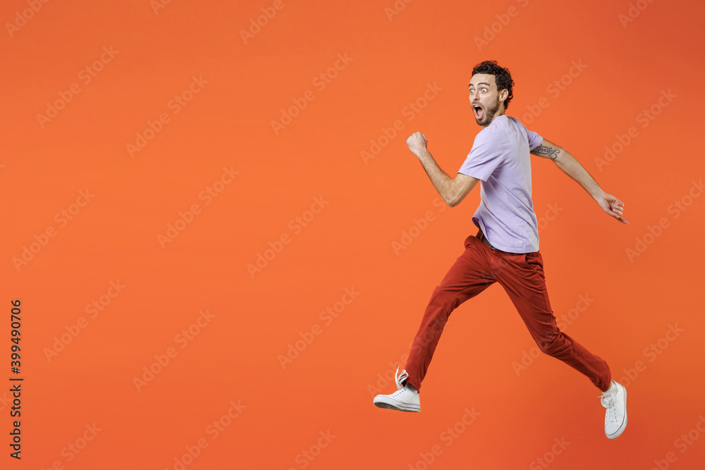 Full length of shocked amazed young bearded man 20s wearing casual violet t-shirt jumping like running keeping mouth open looking aside isolated on bright orange color wall background studio portrait.