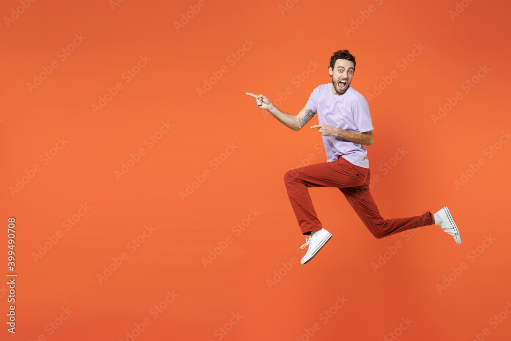 Full length side view of shocked screaming young bearded man 20s wearing casual violet t-shirt jumping pointing index fingers aside isolated on bright orange color wall background studio portrait.