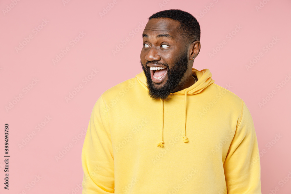 Excited surprised young african american man 20s wearing casual basic yellow streetwear hoodie standing keeping mouth open looking aside isolated on pastel pink color wall background studio portrait. Excited surprised young african american man 20s wearing casual basic yellow streetwear hoodie standing keeping mouth open looking aside isolated on pastel pink color wall background studio portrait.