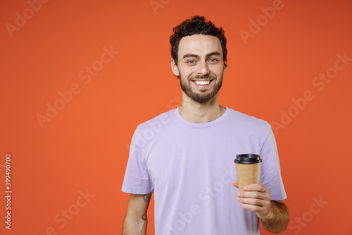 Smiling handsome attractive young bearded man 20s in casual basic violet t-shirt standing hold paper cup of coffee or tea looking camera isolated on bright orange color background studio portrait.