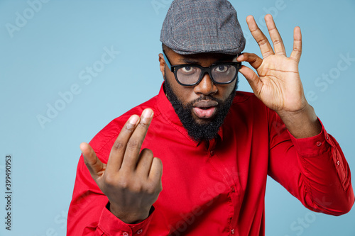 Young bearded african american man 20s wearing casual red shirt cap eyeglasses standing calling with fingers gesture like says come to me isolated on pastel blue color wall background studio portrait.