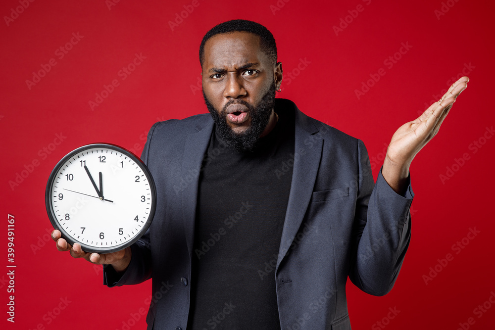 Shocked worried young african american business man 20s wearing classic jacket suit standing spreading hands hold in hand clock looking camera isolated on bright red color background studio portrait. Shocked worried young african american business man 20s wearing classic jacket suit standing spreading hands hold in hand clock looking camera isolated on bright red color background studio portrait.