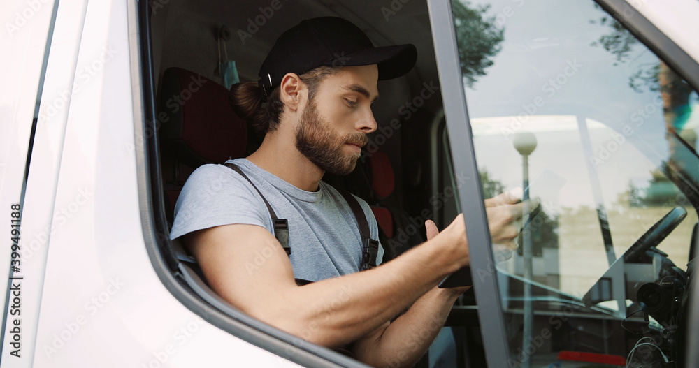 Side view of Caucasian handsome male courier sitting on front seat in van tapping and typing on tablet device delivering orders. Close up of delivery man in uniform using gadget. Work concept Side view of Caucasian handsome male courier sitting on front seat in van tapping and typing on tablet device delivering orders. Close up of delivery man in uniform using gadget. Work concept