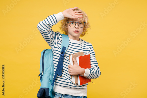 Tired little male kid teen boy 10s years old wearing striped sweatshirt eyeglasses backpack hold school books put hand on head isolated on yellow background child studio portrait. Education concept.