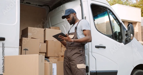Young handsome African American delivery man typing and tapping on tablet device standing near van on street with many ordered boxes. Male postman courier with shopping package. Shipment concept