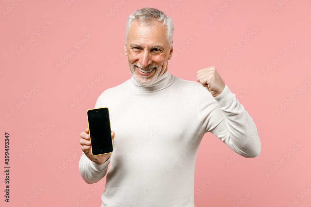 Happy elderly gray-haired mustache bearded man in casual white turtleneck stand doing winner gesture using mobile cell phone with blank empty screen isolated on pastel pink background studio portrait. Happy elderly gray-haired mustache bearded man in casual white turtleneck stand doing winner gesture using mobile cell phone with blank empty screen isolated on pastel pink background studio portrait.