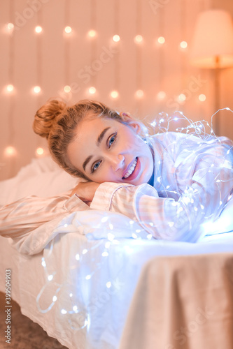 Smiling young woman in pajamas lying on bed with fairy lights, cozy home rela...