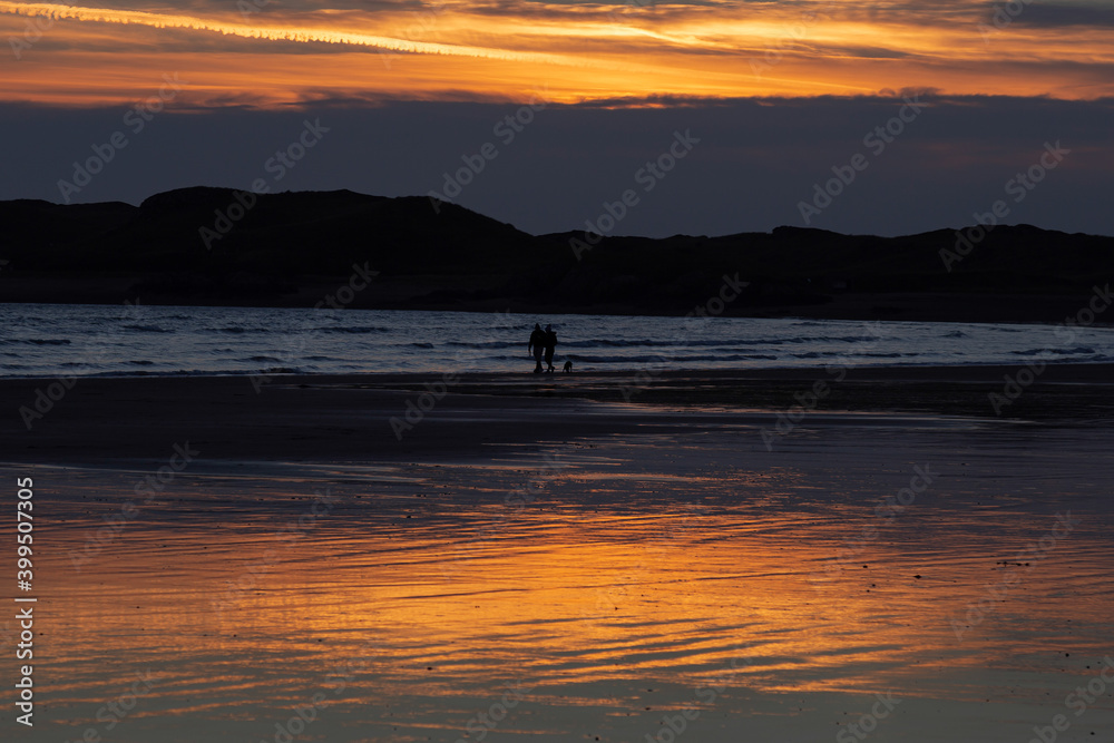 Silhouette of dog walkers on a beach at sunset with hills in the background. Taken at Beaumaris, Anglesey, Wales, with a view across the Menai Straits to the edge of Snowdonia national park.
