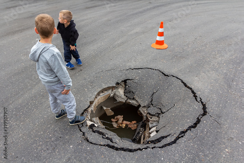 Children play on road near huge deep sinkhole in asphalt surface. Risk of injury to child. Damaged sink hole on city street