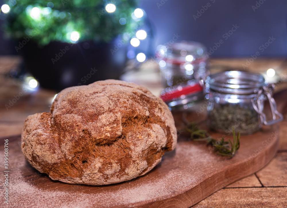 Fototapeta premium Home baked loaf of bread on wooden board, rosemary branch, glass of home-grown spices, Christmas lights soft focus background