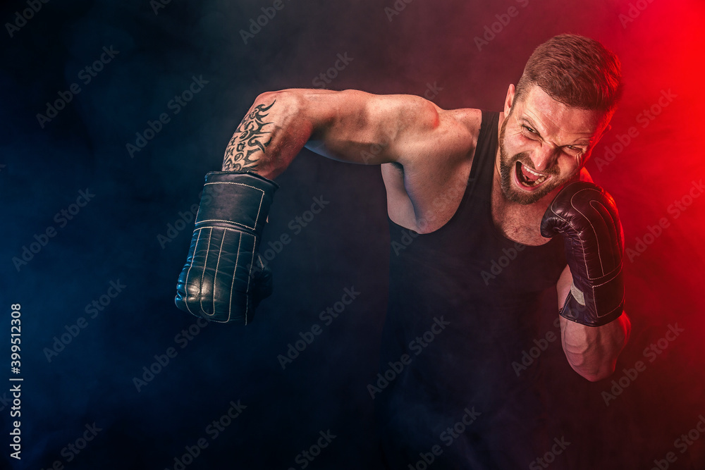 Bearded tattooed sportsman muay thai boxer in black undershirt and boxing gloves fighting on dark background with smoke. Sport concept.