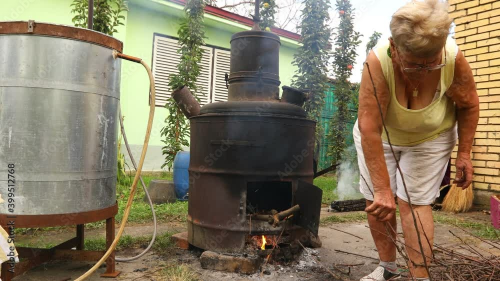 Grandma throws dry branches into the firebox of a homemade distillery making moonshine schnapps, alcoholic beverages Grandma throws dry branches into the firebox of a homemade distillery making moonshine schnapps, alcoholic beverages